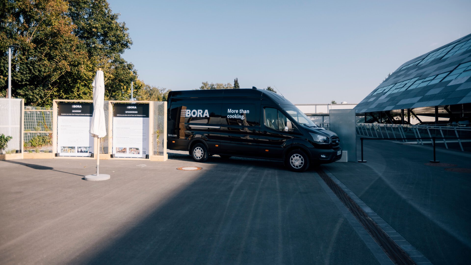 Schwarzer BORA Transporter mit Firmenlogo parkt vor Firmengelände unter blauem Himmel am Tag.