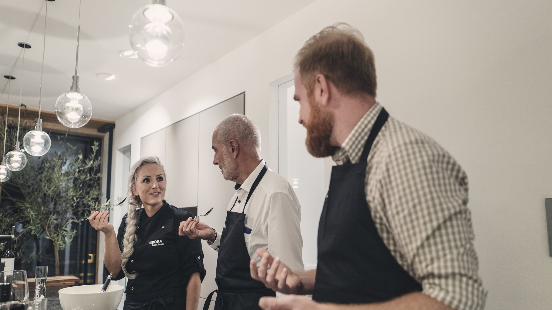 Drei Personen kochen gemeinsam in moderner Küche mit BORA Kochfeldabzug und modernen Pendelleuchten.