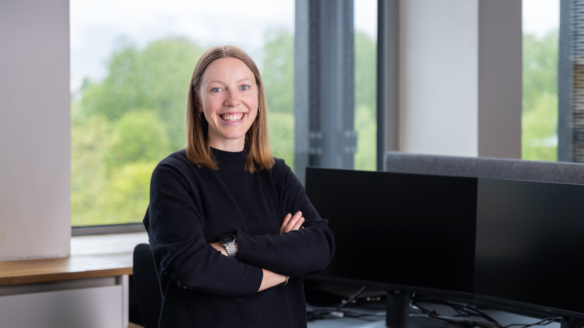 Frau steht selbstbewusst vor BORA Arbeitsplatz mit zwei großen Monitoren und moderner Büroausstattung.
