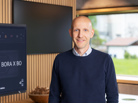 Mann präsentiert BORA X BO Produkt, im Hintergrund BORA Display und moderner Konferenzraum mit Blick ins Grüne.
