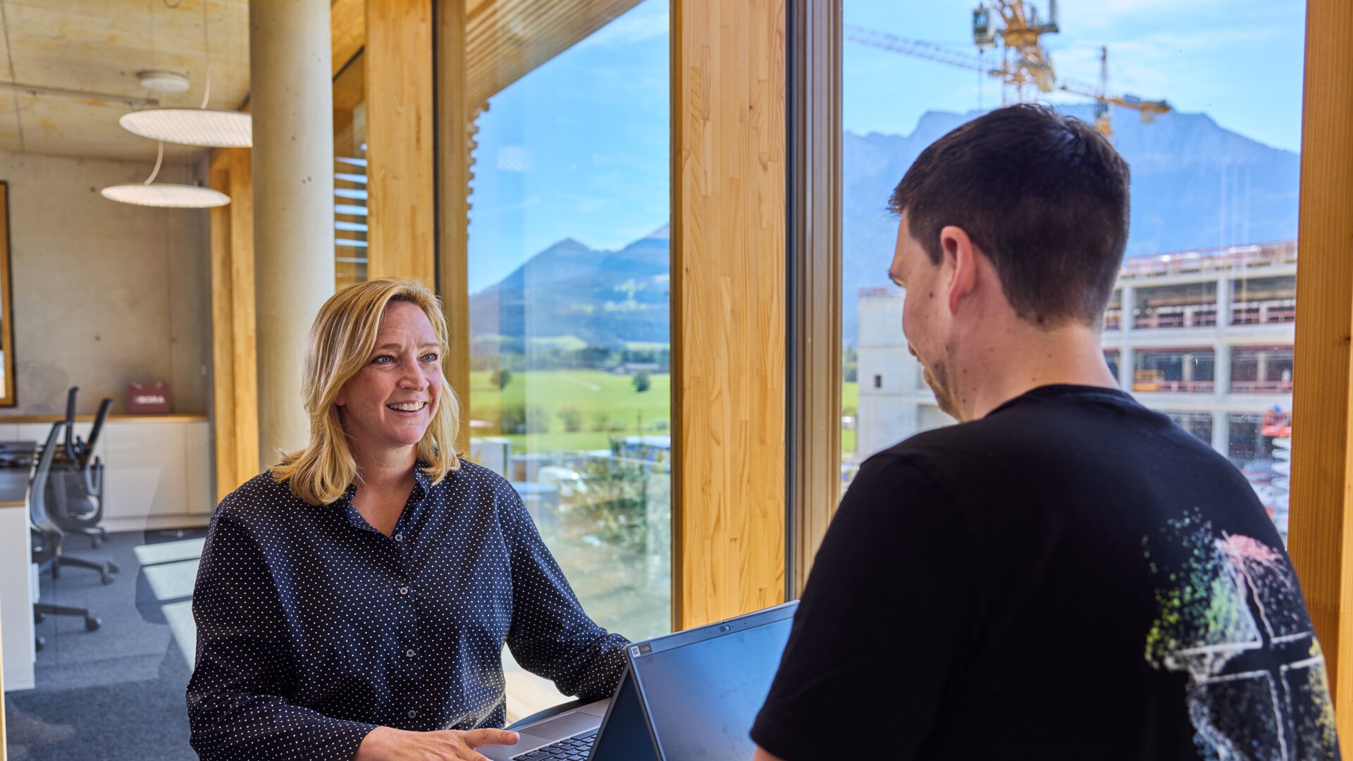 Zwei Personen im BORA Büro arbeiten an Laptops mit Panoramablick auf Berge durch große Fenster.
