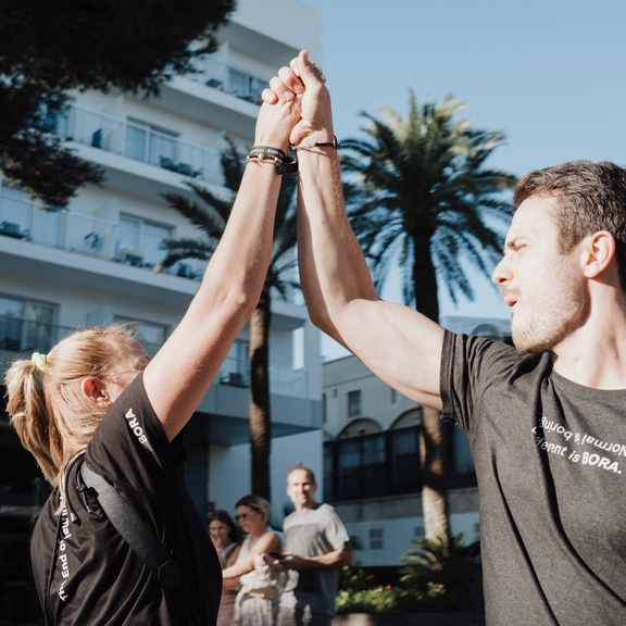 Zwei Menschen mit BORA-Shirts geben sich im Freien einen High Five, im Hintergrund ein Hotel und Palmen.