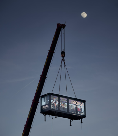 BORA-Werbekabine mit Teamfahrern wird nachts per Kran in die Höhe gehoben, Mond im Hintergrund.