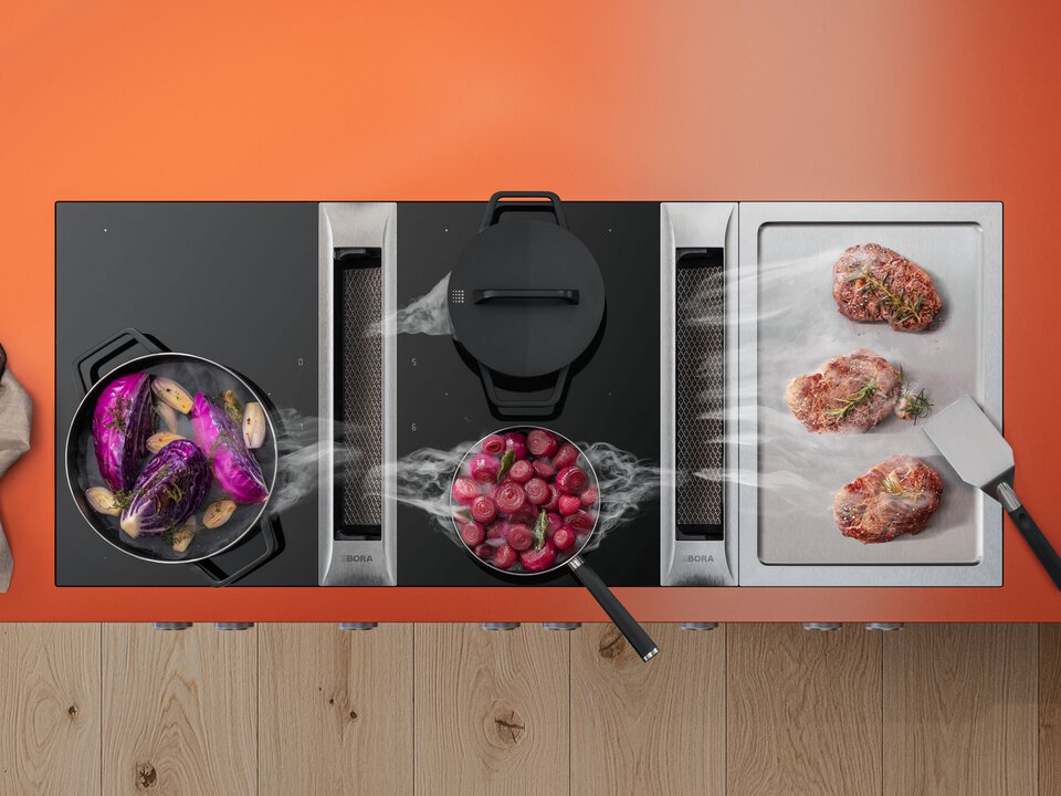 BORA Kochfeld mit integriertem Kochfeldabzug, Dampfabzug über Töpfen und Grillplatte mit Fleisch, moderne Küche in Schwarz und Orange.