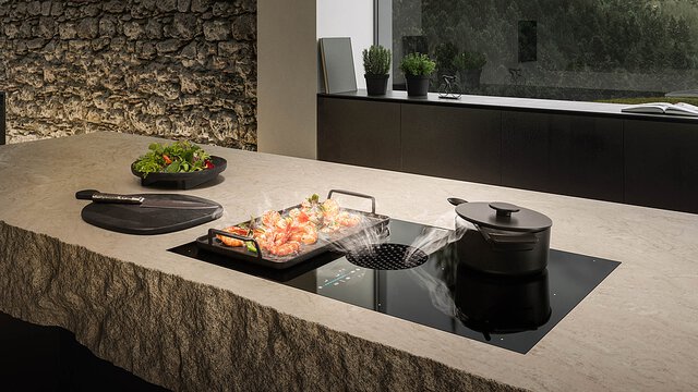 BORA Kochfeld mit integrierter Dunstabzugshaube zeigt frische Garnelen auf Grillplatte und kochenden Topf auf Kücheninsel.