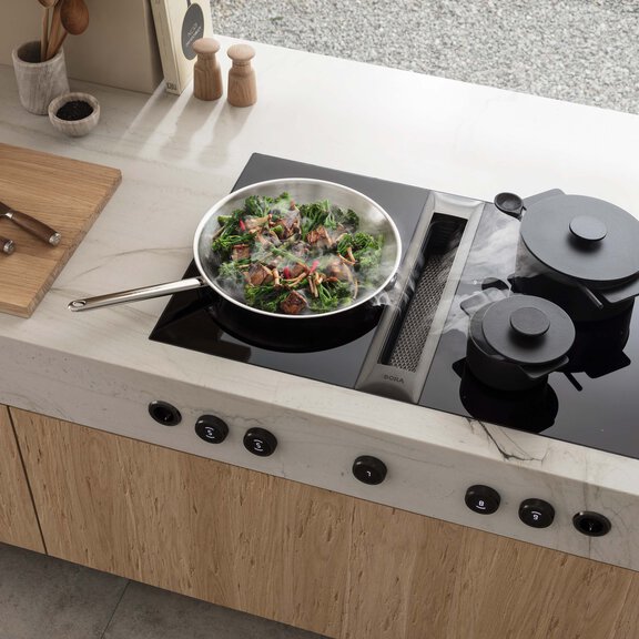 BORA Kochfeld mit integrierter Dunstabzugshaube, zwei Töpfe und Pfanne mit frischem Gemüse auf modernem Küchenblock. BORA Kochfeld mit integrierter Dunstabzugshaube, zwei Töpfe und Pfanne mit frischem Gemüse auf modernem Küchenblock.