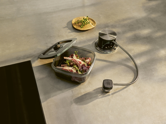 BORA Induktionskochfeld mit passendem Sous-Vide-Garer und Fleisch in vakuumiertem Behälter auf grauer Arbeitsplatte. BORA Induktionskochfeld mit passendem Sous-Vide-Garer und Fleisch in vakuumiertem Behälter auf grauer Arbeitsplatte.