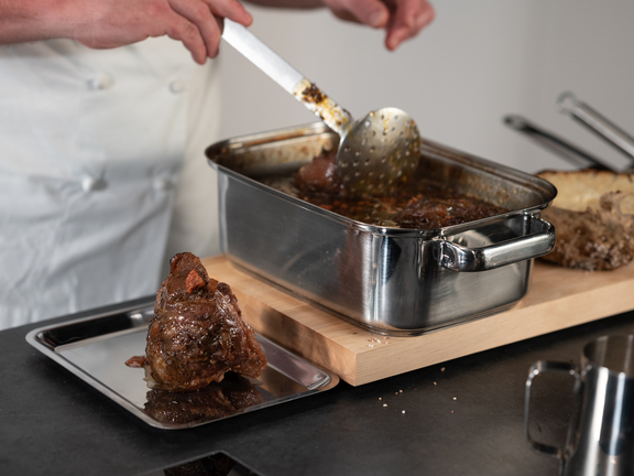 Zubereitung von Fleisch im BORA Kochfeld, Hände eines Kochs entnehmen Braten mit Kelle aus Edelstahl-Bratform.