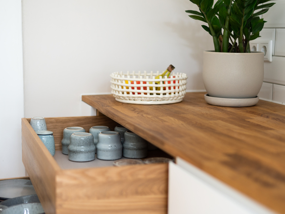 Offene BORA Holzschublade mit grau-blauen Keramikbechern und Holzarbeitsplatte, dekoriert mit Pflanze und Obstkorb. Offene BORA Holzschublade mit grau-blauen Keramikbechern und Holzarbeitsplatte, dekoriert mit Pflanze und Obstkorb.